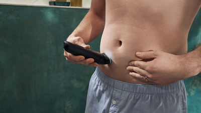 A shirtless man in boxer shorts is using the Philips Bodygroomer to shave his happy trail hair.