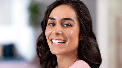 A woman with dark hair and a light pink top is smiling at the camera after finding some sensitive teeth relief.