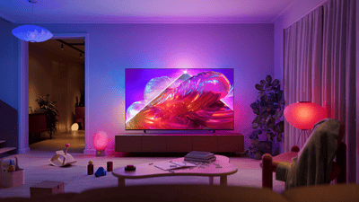 Philips Ambilight TV in a modern living room, displaying vibrant abstract artwork while matching pink, purple, and blue ambient lighting extends onto the wall behind the screen.