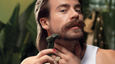 A young man is using a trimmer with a special attachment to trim a scruffy beard.