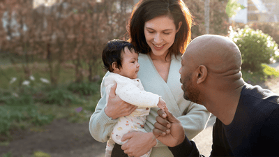 A woman smiles as she holds her baby closely in an outdoor setting. A man reaches out to hold the baby's hand.