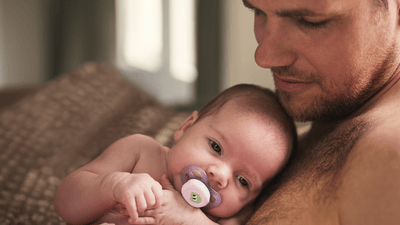 A bare-chested man holds and calms his baby with a Philips Avent Soother, while sitting on a brown sofa.
