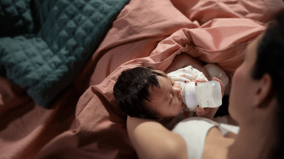 A woman feeds her young infant wrapped in a salmon pink blanket using a Philips Avent baby bottle.