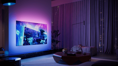 Philips Ambilight TV in a modern living room, displaying a vibrant fantasy scene with immersive blue and purple ambient lighting extending onto the wall behind the screen.
