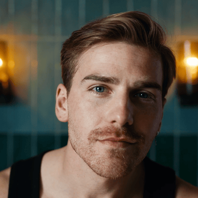A red-haired man with a three-day beard looks into the camera in a bright room.