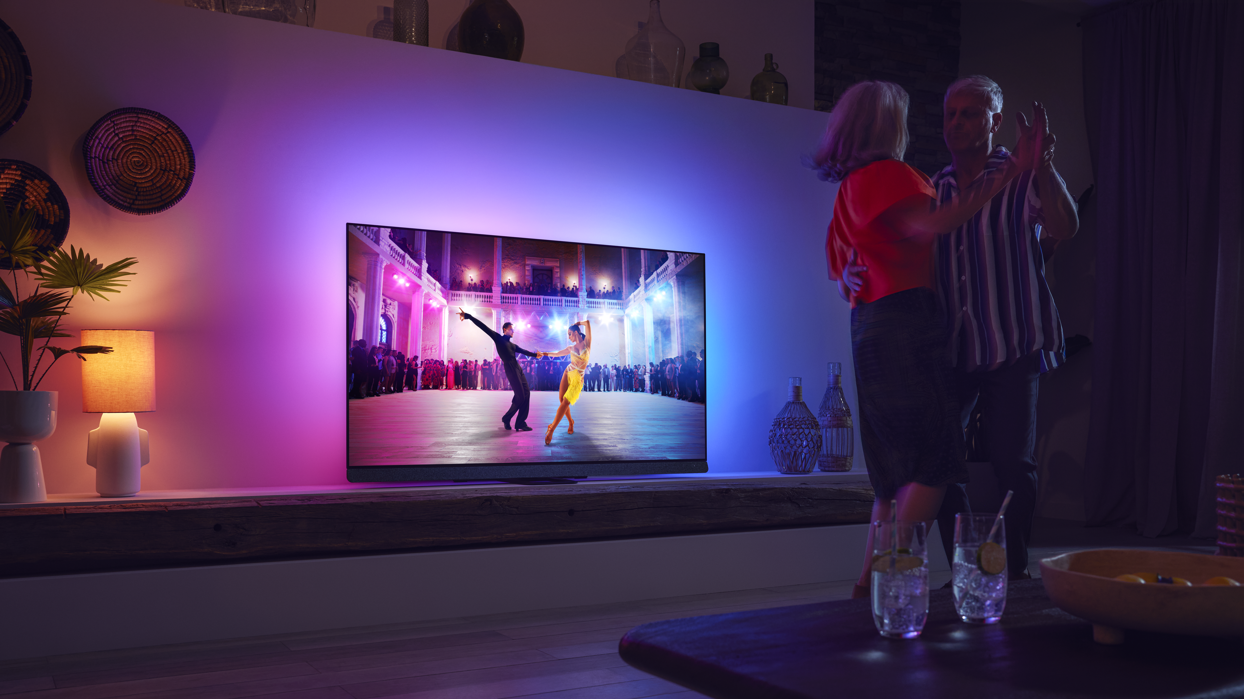 Ζευγάρι που χορεύει στο σαλόνι ενώ παρακολουθεί τηλεόραση Philips Ambilight με πολύχρωμο ατμοσφαιρικό φωτισμό.