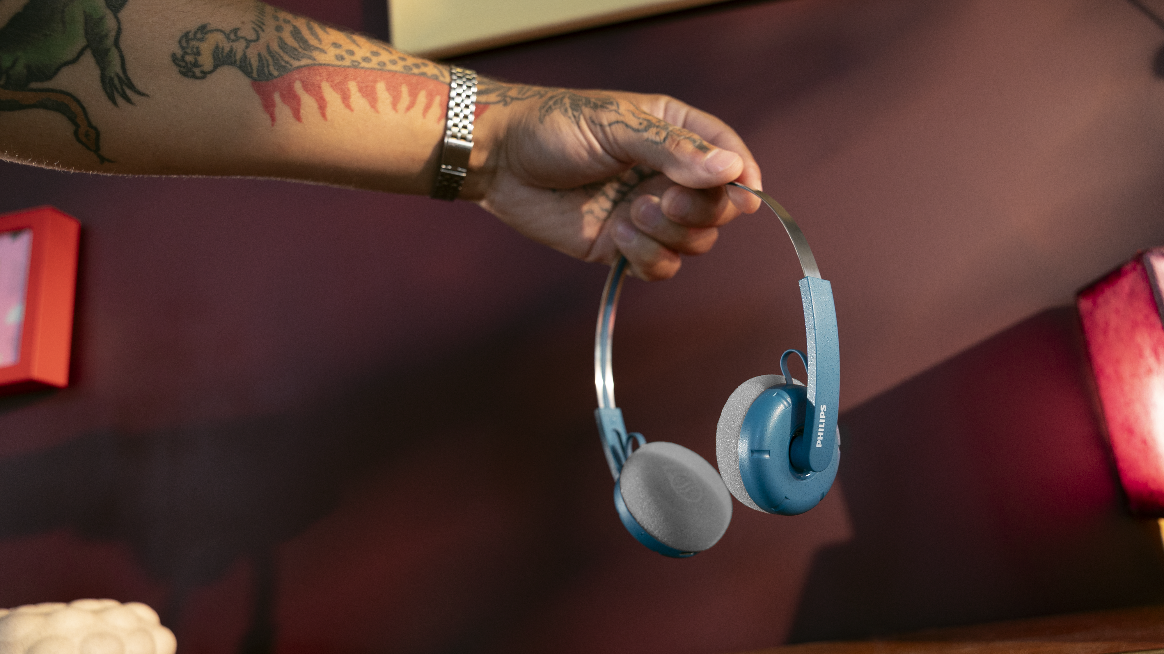 V domačem okolju tetovirana roka drži par modrih naušesnih slušalk Philips nad zofo v pripravi na osebno in pristno izkušnjo poslušanja.