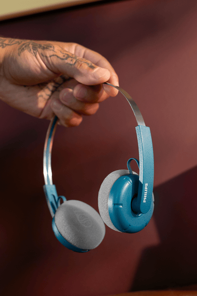 V domačem okolju tetovirana roka drži par modrih naušesnih slušalk Philips nad zofo v pripravi na osebno in pristno izkušnjo poslušanja.