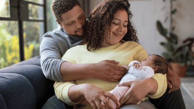 A man wraps his arms around a woman as she holds their sleeping infant while sitting on a blue sofa.