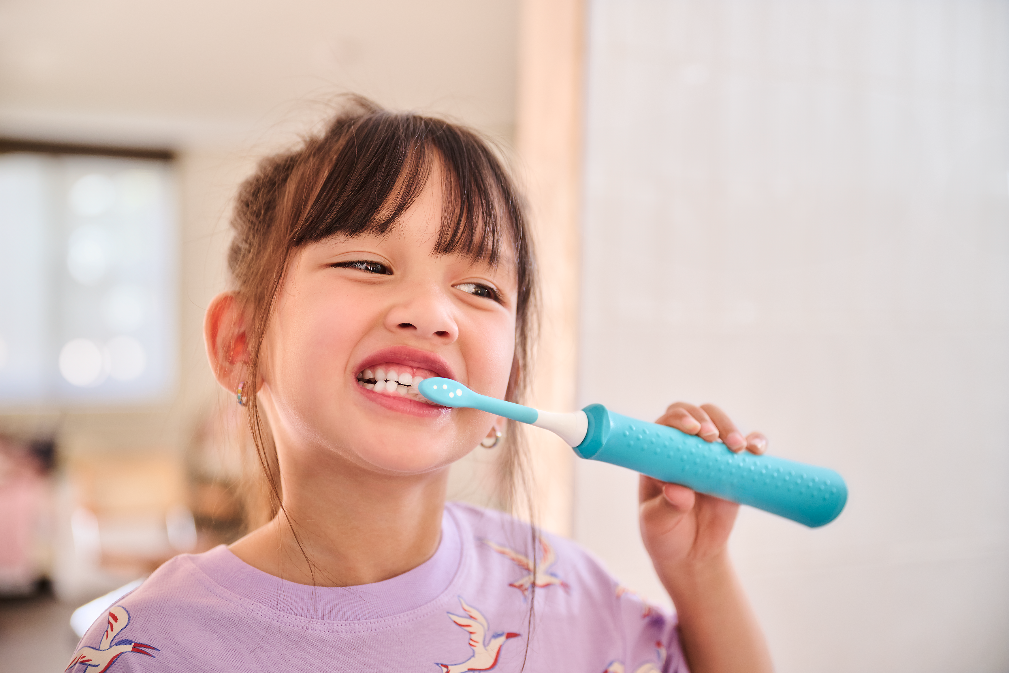 A young child wearing pink is shown brushing her teeth with a blue Philips kids electric toothbrush and kids toothbrush head.