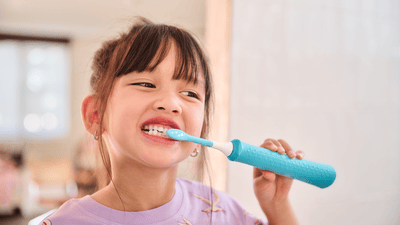 A young child wearing pink is shown brushing her teeth with a blue Philips kids electric toothbrush and kids toothbrush head.