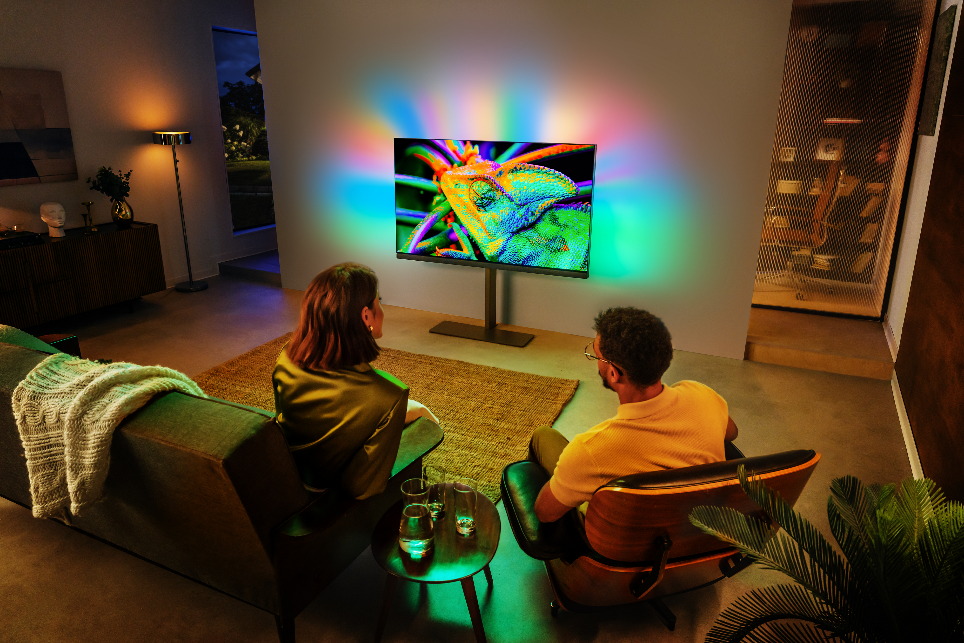 Philips Ambilight‑tv die een kleurrijk beeld toont en omgevingslicht op de muur projecteert terwijl twee mensen in een moderne woonkamer kijken
