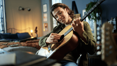 A guy with a cleanly shaved face is smiling in his room while playing the guitar on a chair.