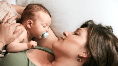 Eine Frau wiegt ihr schlafendes Baby auf einer weißen Bettdecke. Das Baby benutzt einen Philips Avent Schnuller.