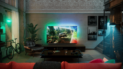 Philips Ambilight TV in a modern loft-style living room, displaying an off-road driving scene with immersive multicolored ambient lighting extending onto the wall behind the screen.