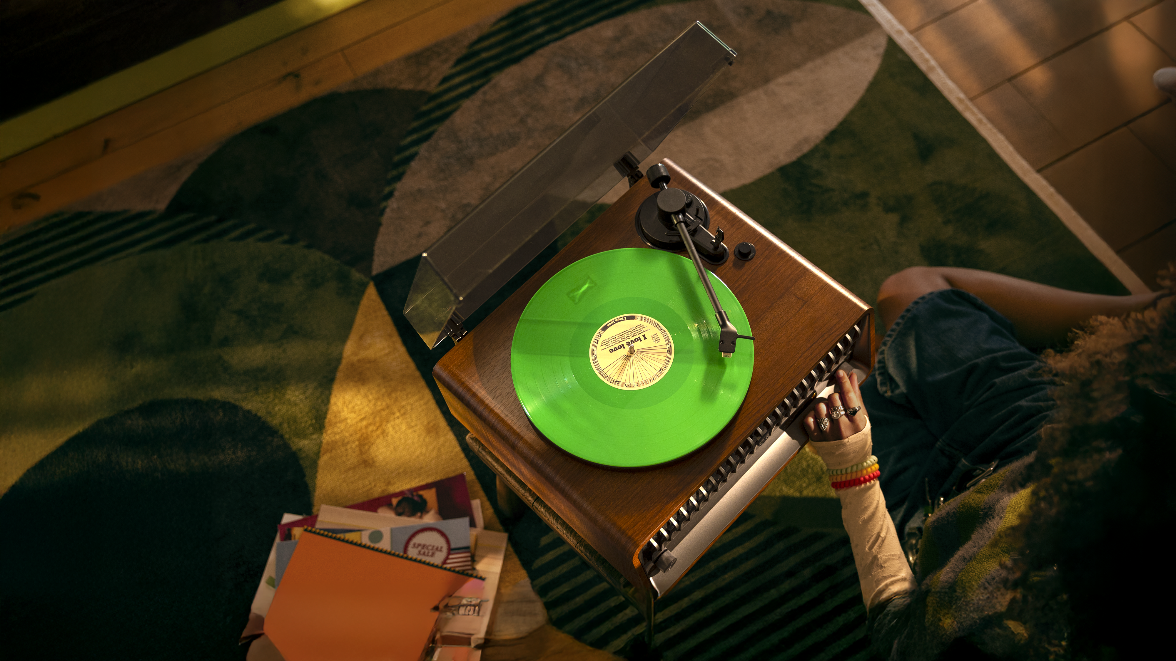 Pogled od zgoraj navzdol na živo zeleno vinilno ploščo, ki se predvaja na lesenem gramofonu v prijetnem domačem okolju. V bližini se sprošča oseba, na mizi pa so raztreseni zvezki, ki poudarjajo izkušnjo Philipsovega zvoka.