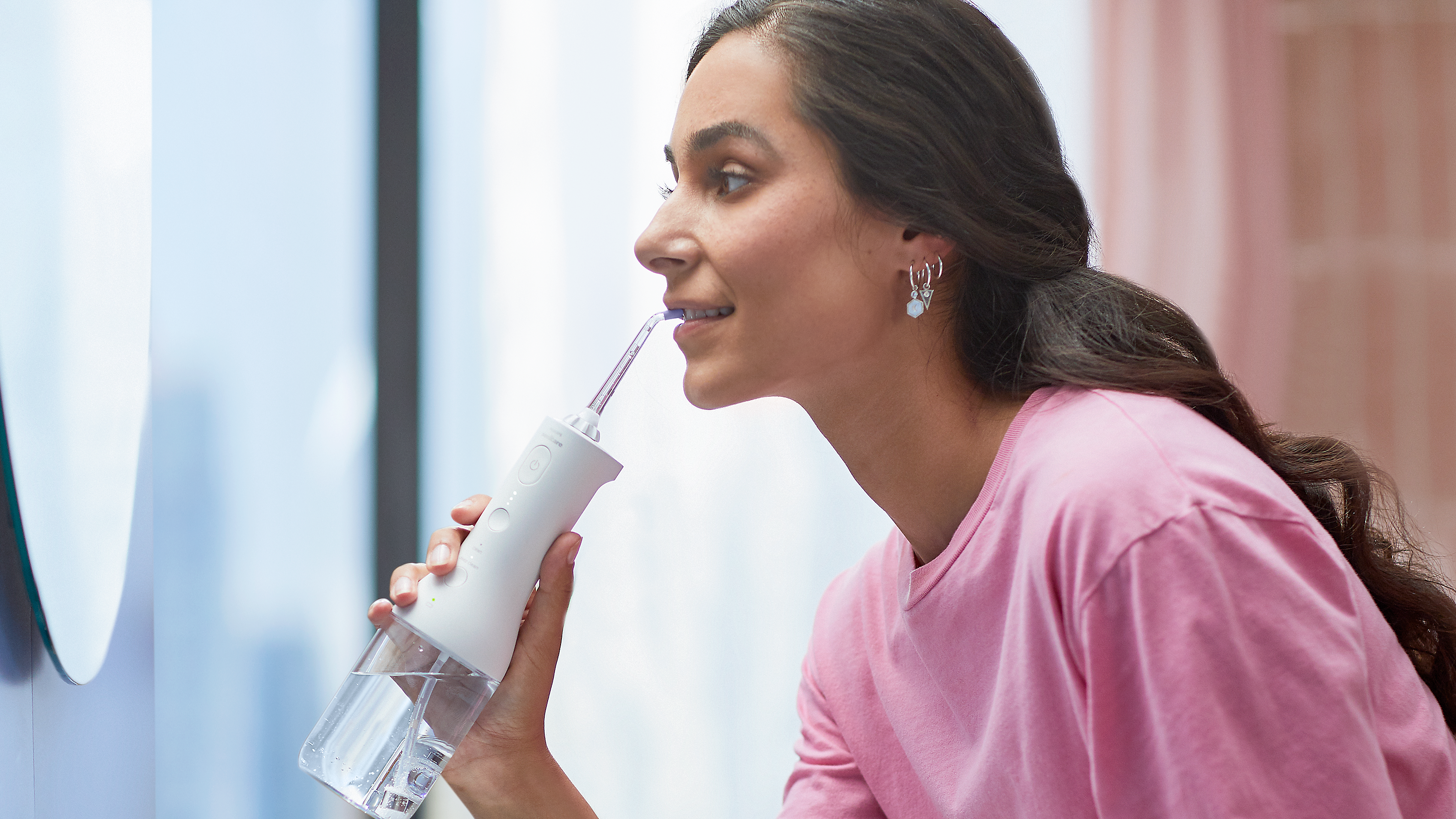 A young woman with long dark hair wearing a pink t-shirt is using the Philips Power Flosser while looking at herself in a mirror.