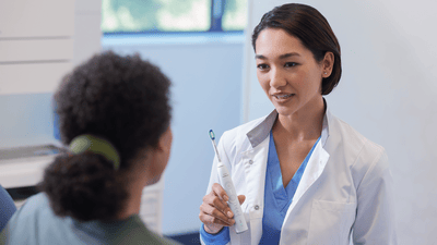 A young dentist professional wearing a white outfit speaks to her patient, holding a toothbrush.