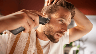 A man is using Philips hair clippers to shave and style the right side of his head into a buzz cut.