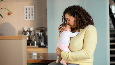 A woman dressed in a light yellow blouse gently kisses her baby's head while sitting beside to a kitchen countertop.