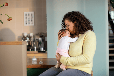 A woman dressed in a light yellow blouse gently kisses her baby's head while sitting beside to a kitchen countertop.