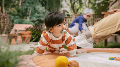 A young child with a soother and in a striped top is crawling on a colourful blanket in a garden.