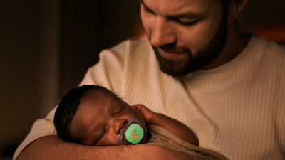 A man gazes down as he holds his sleeping infant in a dimly lit room. The baby is using a Philips Avent Soother.