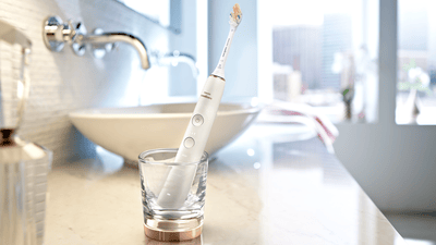 A white Philips electric toothbrush is shown placed upright in a glass tumbler beside a bathroom basin.