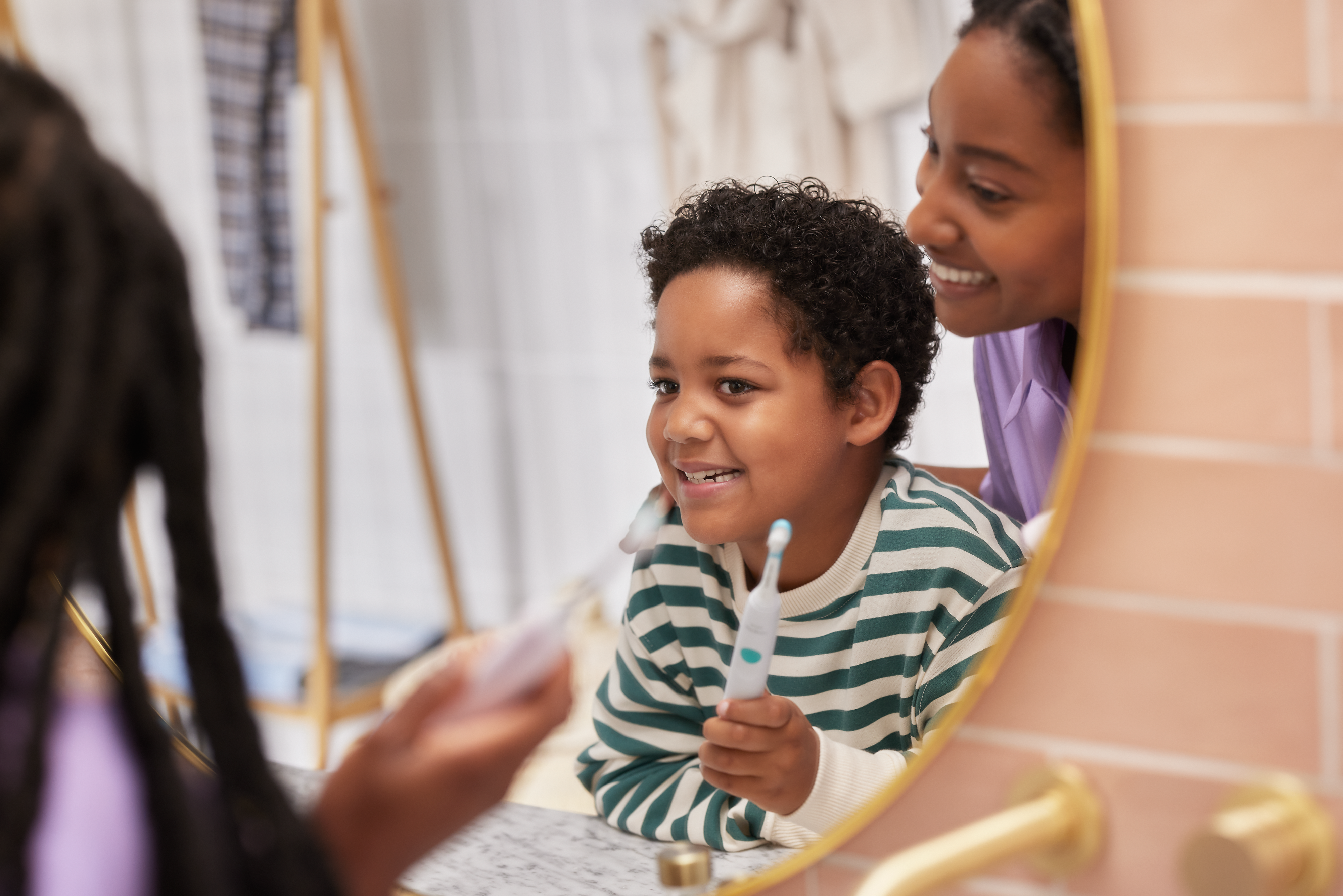 A young mother helps her child to brush their teeth correctly while looking in the mirror.