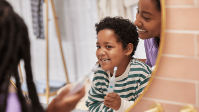 A young mother helps her child to brush their teeth correctly while looking in the mirror.