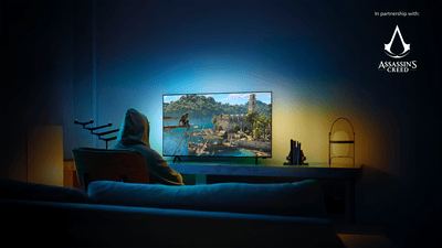 Personne en capuche jouant à Assassin’s Creed sur un TV Philips Ambilight dans une pièce sombre, avec une lumière bleue et ambrée diffusée sur le mur derrière l’écran.