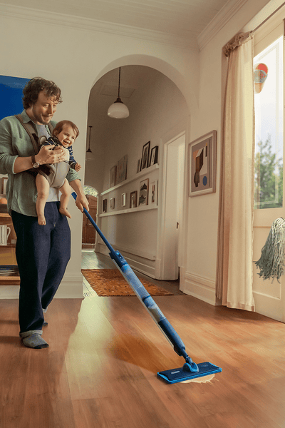 Мъж, държащ бебе, който почиства дървен под с мопа в светла