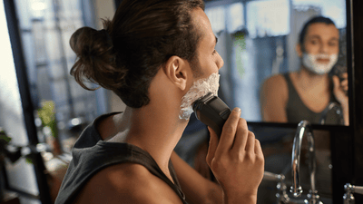 A man with shaving cream on his chin is shaving his beard using a Philips shaver in front of a bathroom mirror.
