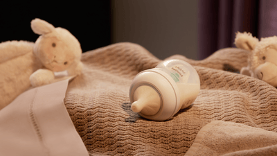 A Philips Avent baby bottle filled with milk lies on its side on top of a brown blanket, surrounded by soft toys.