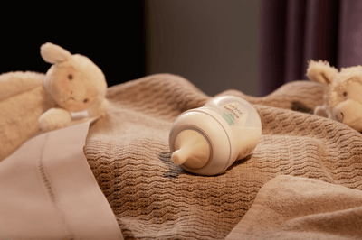 A Philips Avent baby bottle filled with milk lies on its side on top of a brown blanket, surrounded by soft toys.
