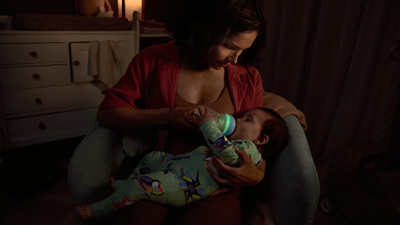 A woman feeds her newborn using a Philips Natural Response Nighttime bottle in a dimly lit bedroom setting.