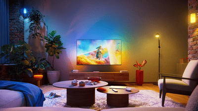 Philips Ambilight TV in a stylish living room, displaying a motocross action scene with colorful ambient lighting extending beyond the screen and illuminating the surrounding wall.