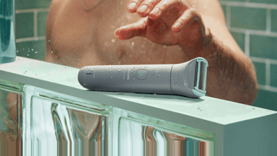 Eine Nahaufnahme eines grauen Philips Bodygroom-Rasierers, der auf einer blauen Theke liegt und von einem duschenden Mann in die Hand genommen werden soll.