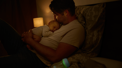 A man gently kisses his sleeping newborn resting on his chest, while a Philips Avent nighttime baby bottle sits nearby.