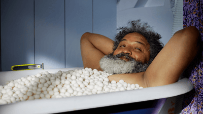 Un hombre con barba poblada se relaja en una bañera llena de guijarros blancos. Cerca hay una Philips OneBlade.