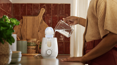 A woman is pouring water from a glass jug into a Philips Avent bottle warmer.