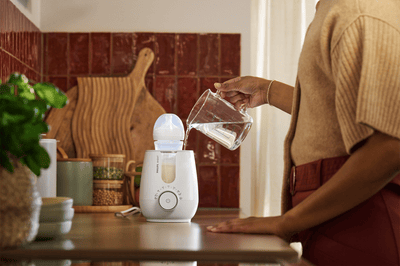 A woman is pouring water from a glass jug into a Philips Avent bottle warmer.