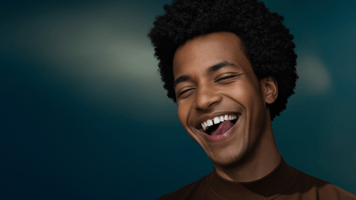 A young man smiles as he licks his white teeth against a dark gradient background.