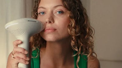 A young woman looking at the camera, using a Philips Lumea 9900 Pro IPL device on her face.