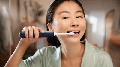 A young woman with dark hair, in a green robe, brushes her teeth with a Philips electric toothbrush.