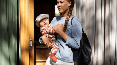A woman holds her baby, who is dressed in a grey hat and striped clothing, on her hip next to a wooden door.