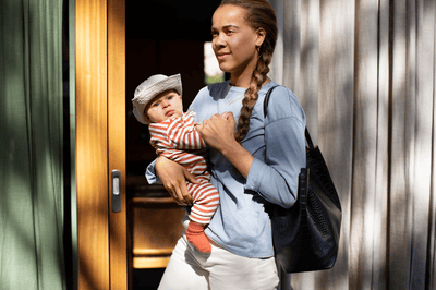 A woman holds her baby, who is dressed in a grey hat and striped clothing, on her hip next to a wooden door.