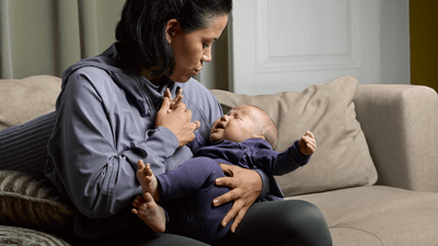 A woman cradles her crying infant on a sofa in a dimly lit living room setting.