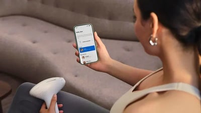 A woman looks at the Philips Lumea IPL app on her phone while using her Philips Lumea 9900 Pro IPL device.