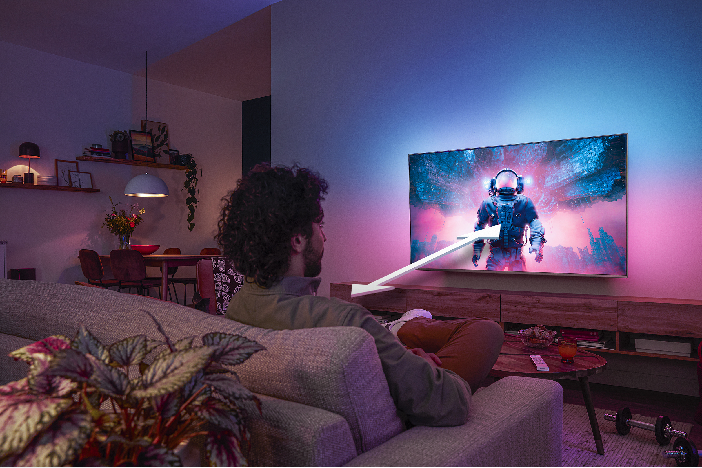 Vyras sėdi ant sofos ir žiūri „Philips Ambilight“ televizorių, kuriame rodomas spalvingas astronauto vaizdas su rodykle, rodančia optimalų žiūrėjimo atstumą